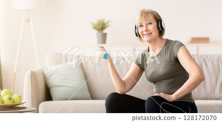 Sporty senior woman sitting on fitball, exercising with dumbbells, listening to music in headphones at home. Beautiful mature lady doing domestic fitness, working out her biceps muscles in living room 128172070