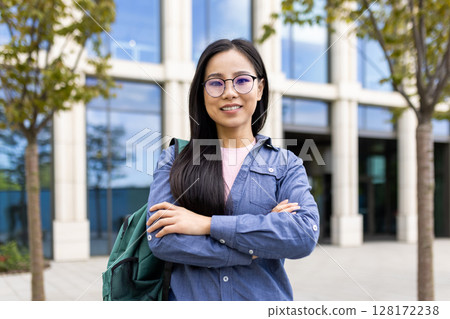 A smiling Asian woman stands confidently with arms crossed, wearing glasses and a backpack in front of a modern building. 128172238