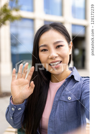An Asian woman waves hello while taking a selfie outdoors, smiling warmly at the camera. An Asian woman waves hello while taking a selfie outdoors, smiling warmly at the camera. 128172239