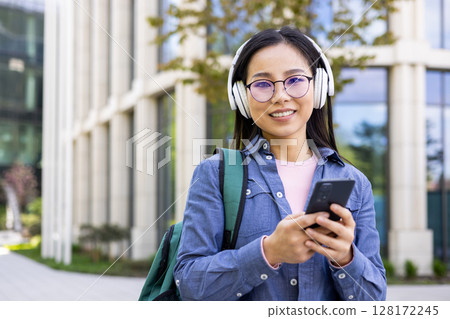 A smiling Asian woman with glasses and headphones holding a phone outdoors near a modern building. A smiling Asian woman with glasses and headphones holding a phone outdoors near a modern building. 128172245