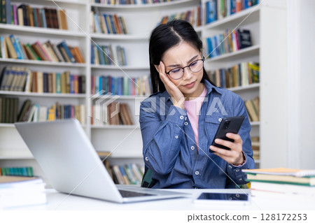 An Asian woman in glasses looks at her phone with a concerned expression in a library, near a laptop. 128172253