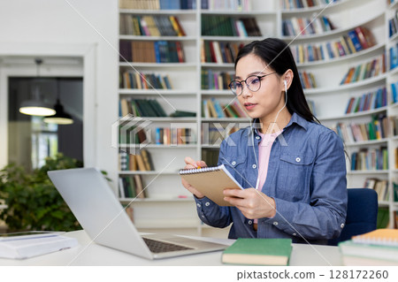 An Asian woman in a library taking notes during a video call, with a laptop and books nearby. An Asian woman in a library taking notes during a video call, with a laptop and books nearby. 128172260