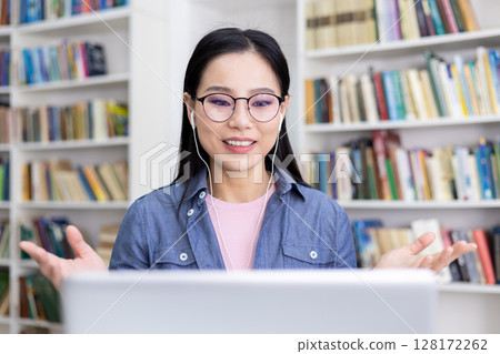 An Asian woman smiles while participating in a video call, using earphones, with a library in the background. An Asian woman smiles while participating in a video call, using earphones, with a library in the background. 128172262