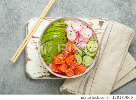 Salmon poke bowl with avocado, radish, cucumber and rice on white rustic wooden board on grey background top view 128173009
