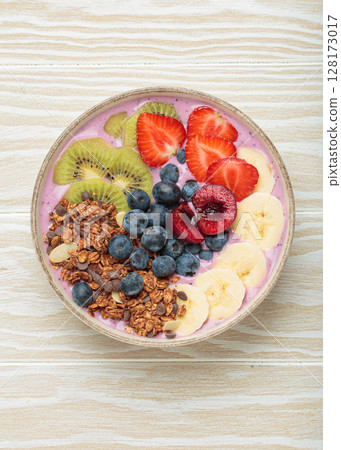 Delicious acai bowl with fresh fruits and granola in white ceramic bowl on white wooden table top view, healthy breakfast or snack 128173017