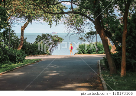 Woman with light backpack walking along green alley in coastal park. Concept of relaxation, sea, travel, active lifestyle, freedom of movement 128173106
