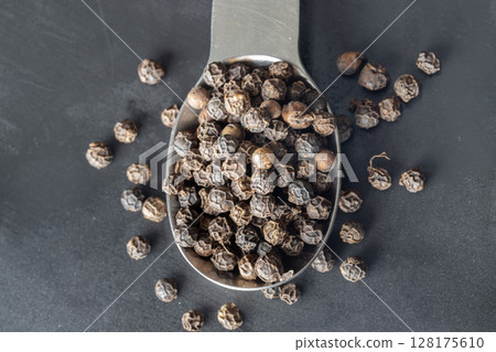 Raw organic black peppercorns or Dry black pepper seeds in stainless steel spoon on dark background. 128175610