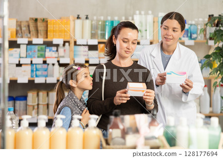 Woman with schoolgirl daughter are busy choosing tablet box at pharmacy, pharmacist in background Woman with schoolgirl daughter are busy choosing tablet box at pharmacy, pharmacist in background 128177694