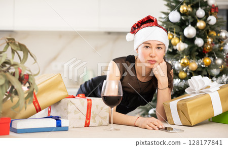 upset young girl in santa hat sits in kitchen. upset young girl in santa hat sits in kitchen. 128177841
