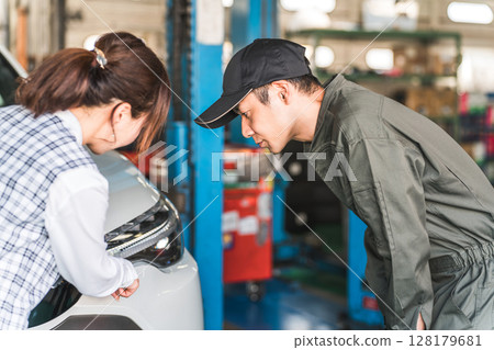 A male car mechanic and a female front desk clerk in office clothes having a conversation in front of a car 128179681