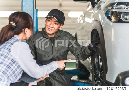 A male car mechanic and a female front desk clerk in office clothes having a conversation in front of a car A male car mechanic and a female front desk clerk in office clothes having a conversation in front of a car 128179685
