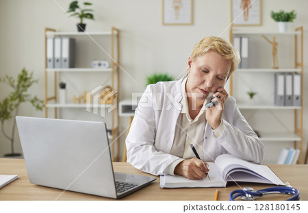 Senior female doctor sitting at desk, talking on mobile phone and taking notes during consultation. Senior female doctor sitting at desk, talking on mobile phone and taking notes during consultation. 128180145