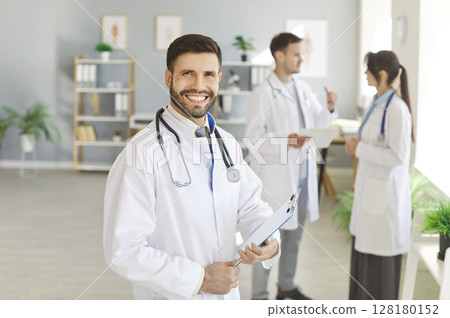 Happy successful young doctor in white uniform looking cheerful at camera and smiling in clinic. 128180152