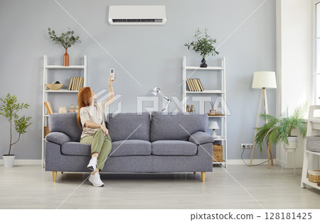 Young woman turning on air conditioner sitting on sofa in the living room at home. 128181425