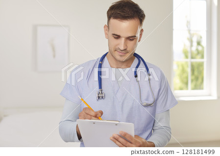 Doctor in light blue medical uniform with stethoscope around his neck is filling out clipboard. Doctor in light blue medical uniform with stethoscope around his neck is filling out clipboard. 128181499