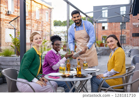 A friendly waiter takes an order from a group of women seated at an outdoor cafe. A friendly waiter takes an order from a group of women seated at an outdoor cafe. 128181710