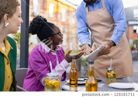 A diverse group of people enjoying a meal outdoors. A waiter takes an order on a tablet. 128181711