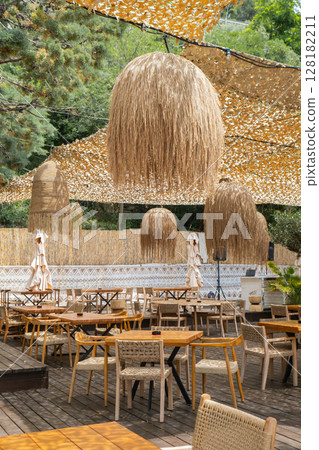 Cozy interior of outdoor cafe on beach seacoast. Wooden tables with thatched bamboo chairs and lamps. Summer terrace with rustic wicker chairs nobody. Natural color rattan furniture restaurant 128182211