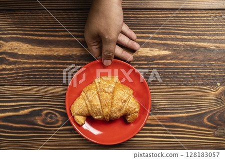 Human hand holding a freshly baked croissant on a red plate with a wooden background. Top view Human hand holding a freshly baked croissant on a red plate with a wooden background. Top view 128183057