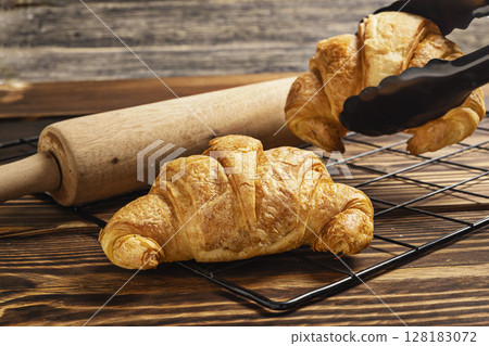 Freshly baked croissants on a cooling tray with tongs and a rolling pin on the table 128183072
