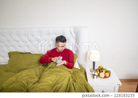 Indonesian southeast asian man sitting on the bed at home gets medicine for coughing. Suffer from fever, flu, and cold. Healthcare and medical concept 128183235
