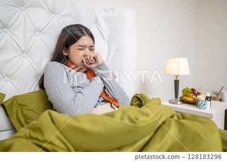 Indonesian southeast asian woman in a blanket sitting on the bed at home and coughing. Suffer from fever, flu, and cold. Healthcare concept Indonesian southeast asian woman in a blanket sitting on the bed at home and coughing. Suffer from fever, flu, and cold. Healthcare concept 128183296