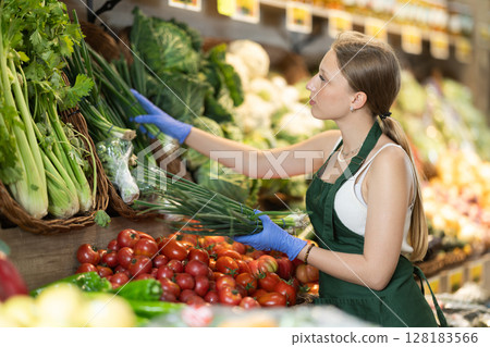 Girl shop seller add bunch of green onion goods on display case in vegetable store Girl shop seller add bunch of green onion goods on display case in vegetable store 128183566