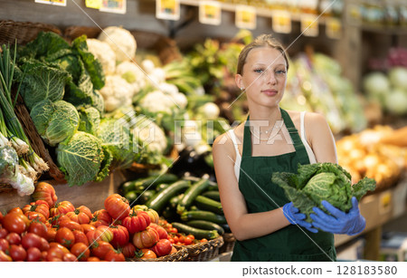 Female grocery store worker offers ripe broccoli cabbage in grocery department of supermarket Female grocery store worker offers ripe broccoli cabbage in grocery department of supermarket 128183580