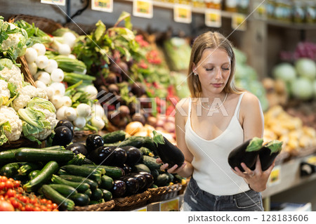 Portrait of female shopper carefully selecting eggplants in supermarket 128183606