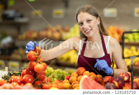 Young female seller puts tomatoes in vegetable shop Young female seller puts tomatoes in vegetable shop 128183710