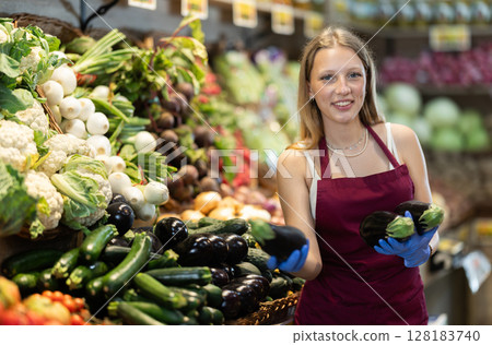 Girl shop seller add eggplant goods on display case in vegetable store Girl shop seller add eggplant goods on display case in vegetable store 128183740