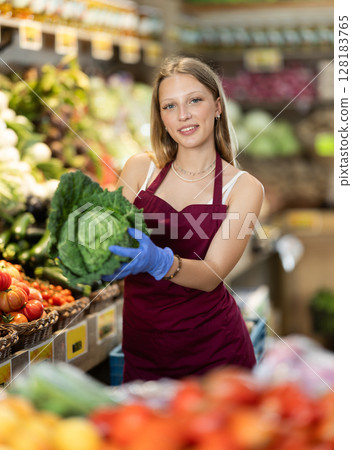 Female grocery store worker offers ripe broccoli cabbage in grocery department of supermarket Female grocery store worker offers ripe broccoli cabbage in grocery department of supermarket 128183765