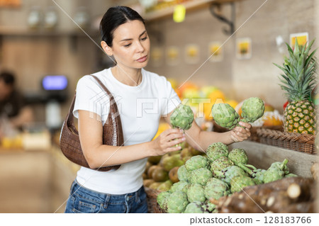 Girl client near vegetable stand in store choose artichoke Girl client near vegetable stand in store choose artichoke 128183766