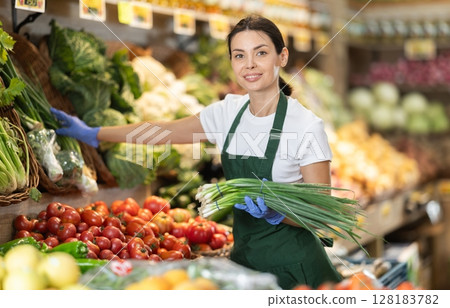 Girl shop seller add bunch of green onion goods on display case in vegetable store Girl shop seller add bunch of green onion goods on display case in vegetable store 128183782