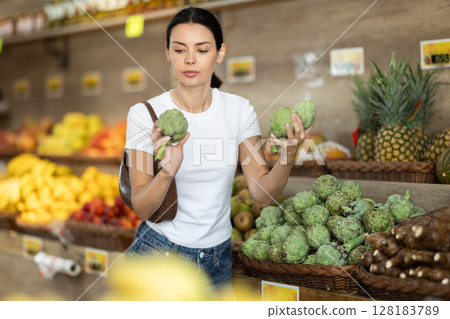 Girl client near vegetable stand in store choose artichoke Girl client near vegetable stand in store choose artichoke 128183789