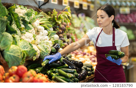 Female salesperson carefully places ripe cucumbers on the counter of grocery supermarket Female salesperson carefully places ripe cucumbers on the counter of grocery supermarket 128183790