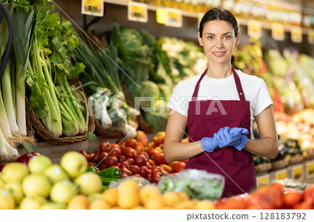 Woman seller in apron and gloves stands in sales area of vegetable store 128183792