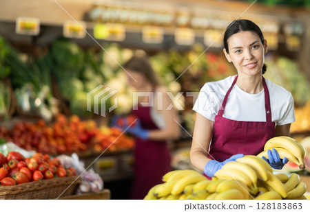 Adult woman seller puts bananas in vegetable shop Adult woman seller puts bananas in vegetable shop 128183863