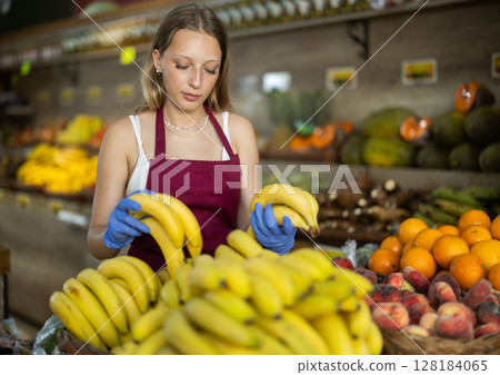 Girl shop seller add banana goods on display case in vegetable store Girl shop seller add banana goods on display case in vegetable store 128184065