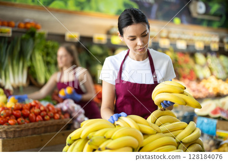Girl shop seller add banana goods on display case in vegetable store 128184076
