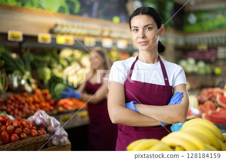 Woman seller in apron and gloves stands in sales area of vegetable store 128184159