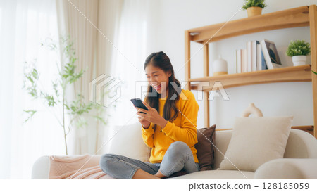 Happy young asian woman relax on comfortable couch at home texting messaging on smartphone 128185059