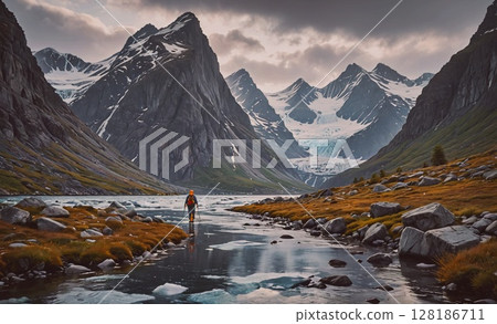 Man is walking along a river in the mountains. The mountains are covered in snow and the sky is cloudy. The scene is peaceful and serene, with the man enjoying the beauty of nature 128186711
