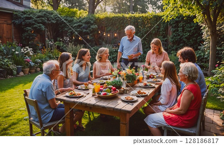 Family is gathered around a wooden dining table in a backyard, enjoying a meal together. The atmosphere is warm and inviting, with everyone smiling and laughing. The table is filled with various 128186817