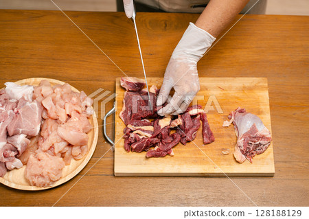 Asian man's hands cutting meat on a wooden cutting board in the kitchen, preparing to cook. 128188129