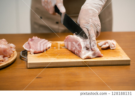 Asian man's hands cutting meat on a wooden cutting board in the kitchen, preparing to cook. 128188131