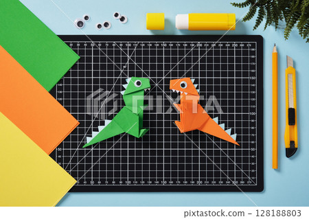 Green and orange origami dinosaurs on a black cutting mat. Surrounded by colorful sheets of paper, googly eyes, glue stick, pencil, and craft knife. DIY project concept for kids. 128188803
