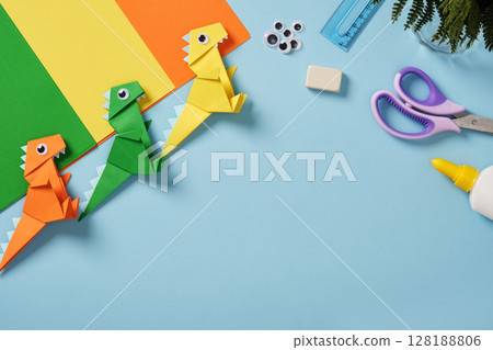 Origami work table with paper, googly eyes, scissors, glue bottle, ruler, and eraser on a light blue background. Finished paper dinosaur toys. Space for your text. Origami work table with paper, googly eyes, scissors, glue bottle, ruler, and eraser on a light blue background. Finished paper dinosaur toys. Space for your text. 128188806