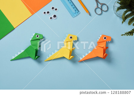 Origami work table with sheets of paper, googly eyes, scissors, ruler, and eraser on a light blue background. Finished paper dinosaur toys. Origami work table with sheets of paper, googly eyes, scissors, ruler, and eraser on a light blue background. Finished paper dinosaur toys. 128188807