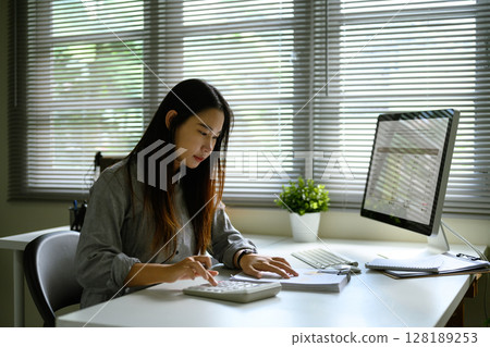 Close up of woman using a calculator and reviewing financial reports at her desk Close up of woman using a calculator and reviewing financial reports at her desk 128189253